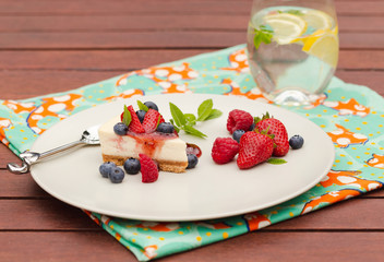 Cheesecake with berries, coulis and mint with glass of iced water and lemon on wooden table with green napkin