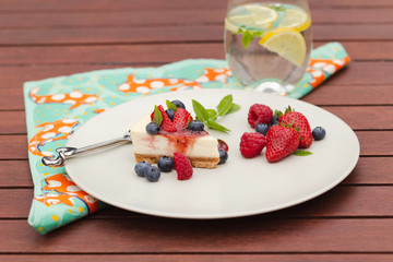 Cheesecake with berries, fruit coulis and mint with glass of iced water and lemon on wooden table with bright green and orange napkin