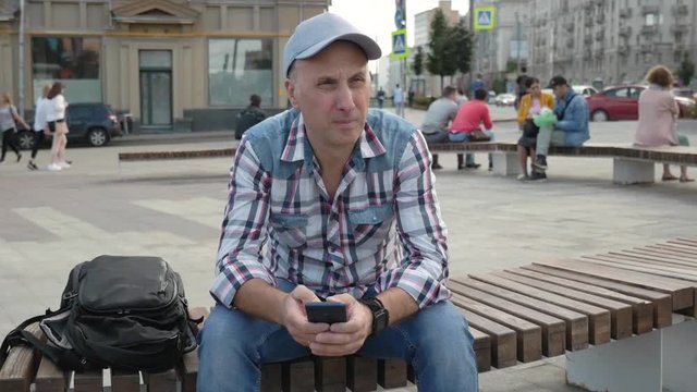 kaukasian gloomy middle-aged man gray plaid shirt jeans black backpack sitting on bench in squarecity sends an SMS message on a smartphone Summer sunny day People go Cars passing Teens on skateboard