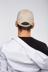 Cropped back view shot of a dark-haired man, wearing beige baseball cap, black t-shirt and white shirt. He is posing on grey background. 