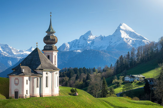 Classic panoramic view of scenic snow-capped Watzmann mountain top with historic pilgrimage church Maria Gern on a beautiful sunny day in spring, Nationalpark Berchtesgadener Land, Bavaria, Germany