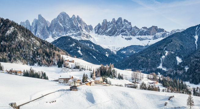 Classic panoramic view of famous Dolomites mountain peaks with the idyllic village Val di Funes and historic Church of St. Magdalena on a scenic sunny day with blue sky in winter, South Tyrol, Italy