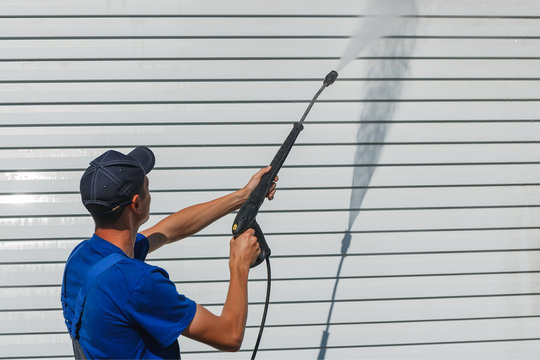 Worker in overalls washes a white wall from a siding with a water gun
