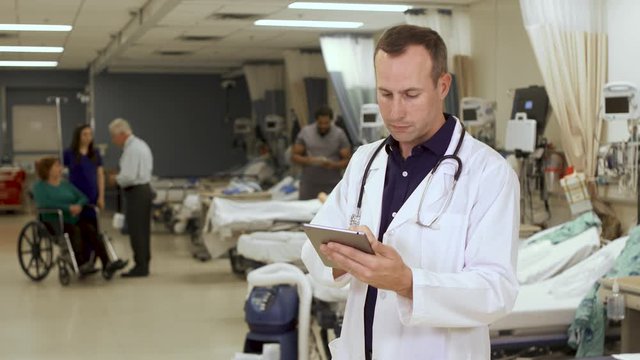 Tech Savvy Doctor Uses A Tablet Computer In A Hospital With Nurses And Patient In A Wheelchair In The Background.