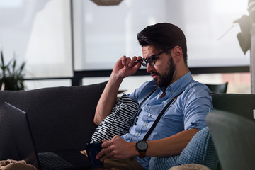 Fototapeta premium Young bearded businessman working on laptop computer while sitting on sofa at his home office