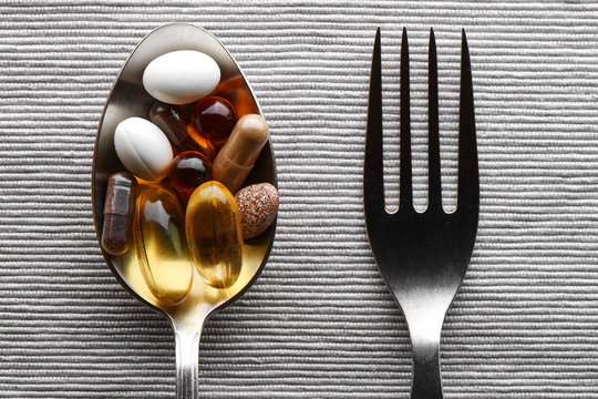 Close-up of a tablespoon and fork filled with various dietary supplements, tablets and vitamins on a gray fabric background