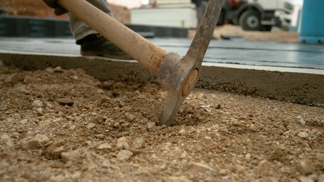 SLOW MOTION, CLOSE UP, DOF: Metal pickaxe strikes into the soft soil mixed with gravel. Unrecognizable worker uses a pickaxe to start digging into the gravel ground next to house under construction.