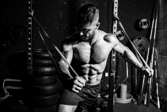 Young sweaty strong focused fit muscular man chest stretching cross workout training in improvised gym with rubber for strength and good looking of muscles dark image black and white