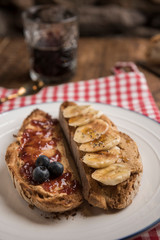 Vegan breakfast, toasted country loaf with peanut butter, banana, blueberries, cinnamon, maple syrup and strawberry jam. Black coffee in the background. Vertical