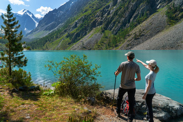 Young tourists with backpacks on the shore of the lake with turquoise water enjoy the view of the mountain peaks.