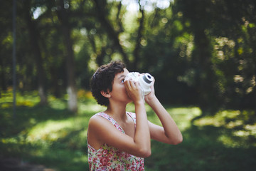 Enthusiastic preteen girl raising instant camera up and taking photo in green park