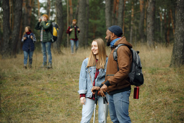Fototapeta premium Group of friends on a camping or hiking trip in autumn day. Men and women with touristic backpacks going throught the forest, talking, laughting. Leisure activity, friendship, weekend.