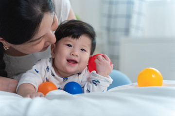 Fototapeta na wymiar Young beautiful asian mother with asian baby on bed and playing toy ball together on white bed with feeling happy and cheerful and the baby that crawling on the bed.Baby family concept