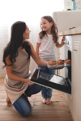 mother looking at daughter taking muffins from the oven