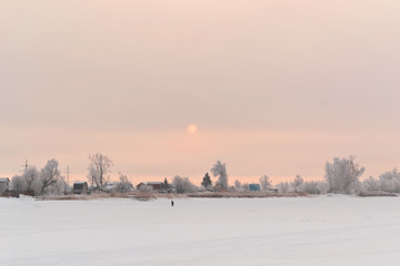 Natural snow-covered landscape with rising sun in frost, winter at dawn. A fisherman walks on a frozen river in the distance.