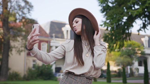 Attractive portrait of charming happy cheerful lady in trendy hat and clothes which making funny selfie on her smartphone during walk near luxurious dwellings