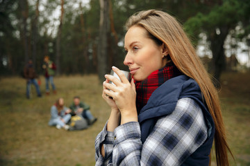 Close up of woman drinking coffee or tea with group of friends on a hiking trip in autumn day. People with touristic bags in the forest, talking, laughting. Leisure activity, friendship, weekend.
