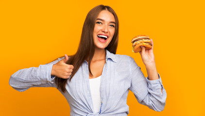 Girl Holding Burger Gesturing Thumbs-Up Standing On Yellow Background