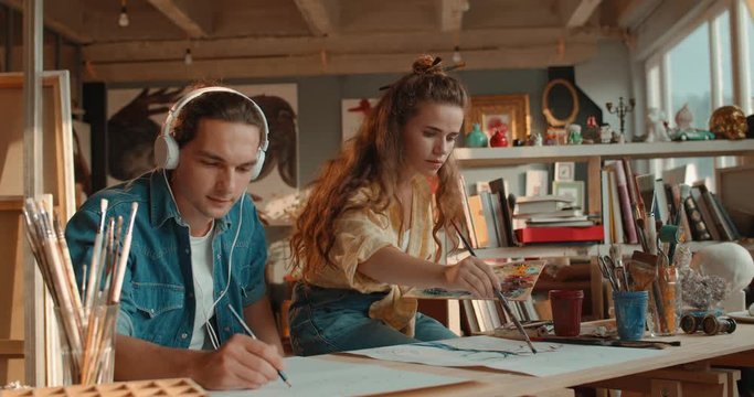 Caucasian young pretty woman painting a picture with paints and handsome man in headphones listening to the music and drawing with a pencil on the white sheet of paper.