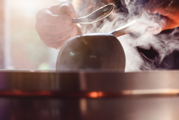 Cooking person in the restaurant is cooking while using the dipper in a large pot. And the water is boiling, there is smoke, hot spit.