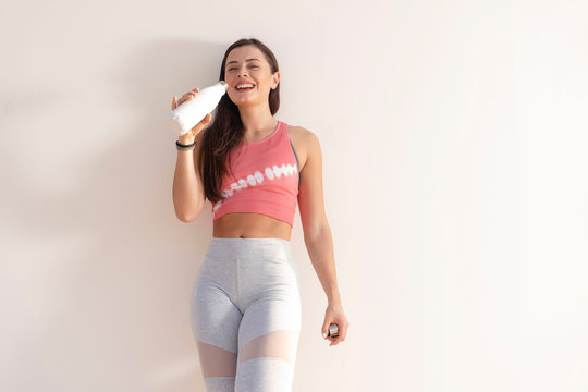 Beautiful Happy Brunette Fitness Model And Wearing A Pink Crop Top And Gray Yoga Tights Standing Against A Plain Beige Wall With A White Water Bottle