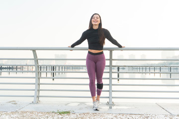Beautiful brunette fitness model facing camera wearing a black long sleeve top and purple tights pose on a fence with a cityscape background on a bright sunny day
