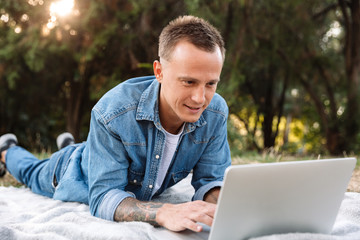 Photo of man lying on blanket in park and typing on laptop computer