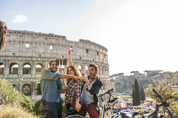 Three happy young friends tourists with bikes at Colosseum in Rome taking pictures and selfies with...