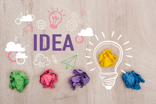 top view of multicolored crumpled paper balls near idea word, light bulb and multimedia icons illustration on wooden surface, business concept