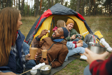 Group of friends on a camping or hiking trip in autumn day. Men and women with touristic backpacks having break in the forest, talking, laughting. Leisure activity, friendship, weekend. Eat and drink.