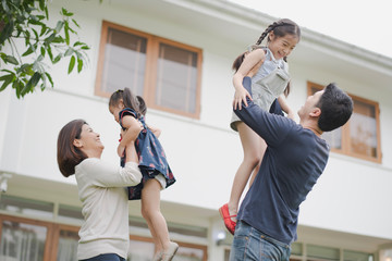 Young Asian family love, father mother and daughter standing playing and fun at outdoor in front of home. parent lift up daughter which smiling and felling happy. background is white house.