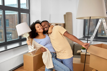 Fototapeta na wymiar moving, people, repair and real estate concept - happy african american couple with lamps packing stuff into cardboard boxes at home and hugging