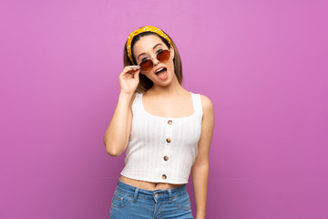 Pretty young woman over isolated purple wall with glasses and surprised