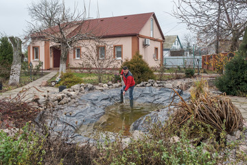 Fototapeta na wymiar White middle-aged man cleans a garden pond with landing net from slime, water plants, falling leaves and catches fish.