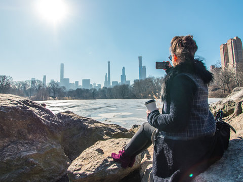 Young woman sitting on rocks by frozen lake in Central Park taking pictures of Manhattan