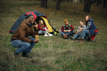 Group of friends on a camping or hiking trip in autumn day. Men and women with touristic bag having break in the forest, talking, laughting. Leisure activity, friendship, weekend. Drink in thermos.