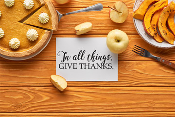 top view of delicious pumpkin pie, apples and card with in all things give thanks illustration on wooden orange table
