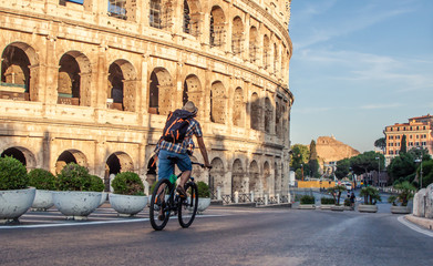 Happy young man tourist with bike wearing shirt and hat at colosseum in Rome, Italy at sunrise.