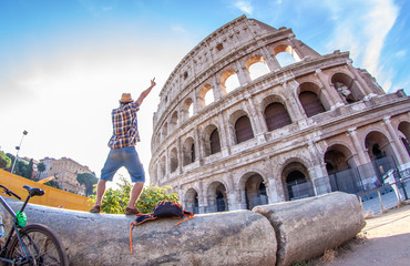 Happy young man tourist with bike wearing shirt and hat standing on a column taking pictures at colosseum in Rome, Italy at sunrise.