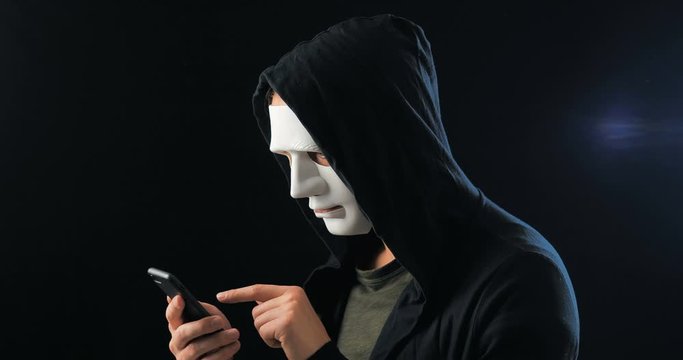 Anonymous masked fraudster in hood demands a ransom for blackmail using a smartphone. Masked Criminal intimidates the victim with the help of threats via SMS using mobile phone.