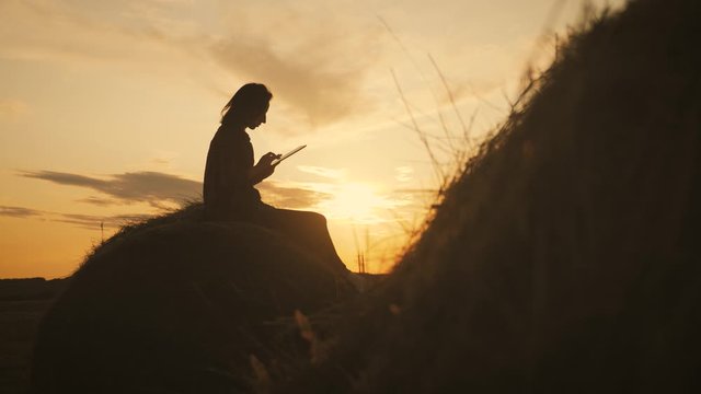 Girl farmer uses tablet in the field next to haystack at sunset. Smart farming, using modern technologies in agriculture.