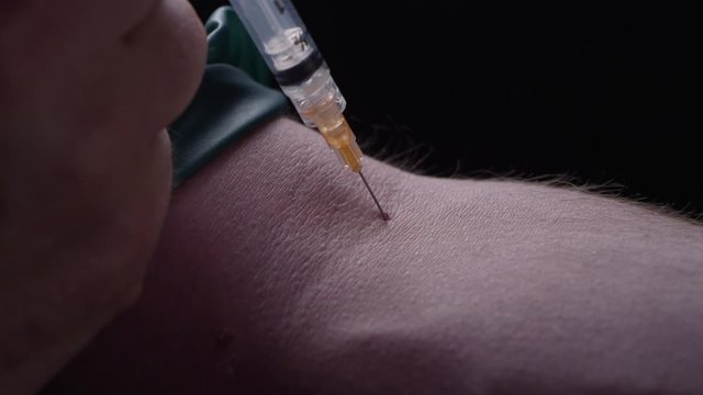 Close-up of a man puncturing his skin with a needle to shoot up heroin, blood leaks from the wound