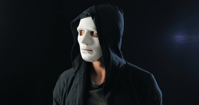 Anonymous man in a white mask covering his face and a black hood turns and looks at the camera. Black dark background. Concept of a member of a secret organization