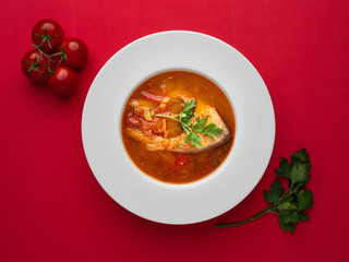 Fish soup in white plate with fresh tomatoes and parsley on red background. Overhead view.