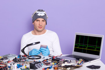 Young computer specialist repairing broken smart phone, being ready to dismantle it, siiting at table full of tools, radiotrician testing electronic equipment in service centre. Electronic engineering