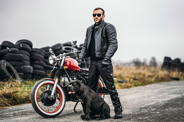 Biker in a leather suit and his dog stand near a red motorcycle on the road. Many tires on the...