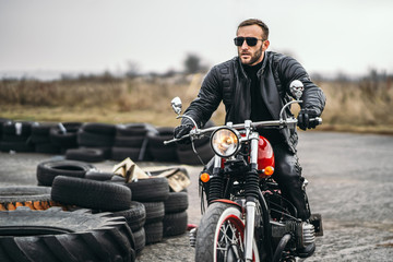 Bearded man in sunglasses and leather jacket sitting on a red motorcycle and looking on the road....