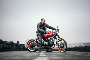 Fototapeta na wymiar Red motorbike with rider. A man in a black leather jacket and pants stands sideways in the middle of the road. Tires are laid on the background