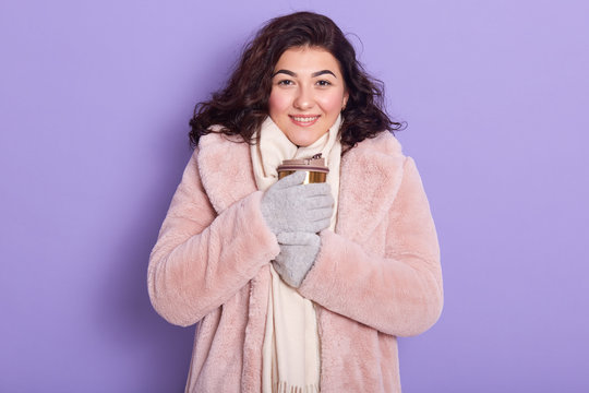 Wimsome freezing young woman drinking hot beverage,trying to bask her hands, holding thermo mug, female dresses pale pink faux fur coat and white scarf, having dark wavy hair. Cold weather concept. - Powered by Adobe