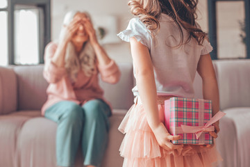 girl holding present box in the hands behind her back and making surprise for grandmother sitting on the couch